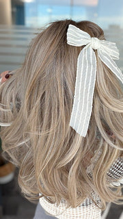 Roxie wearing BROOKLYN by BELLE TRESS in color MILKSHAKE BLONDE R | Mixture of Light sandy brown, highlighted with light ash blonde with a hint of light platinum blonde with medium root color. (styled with bow - not included)
